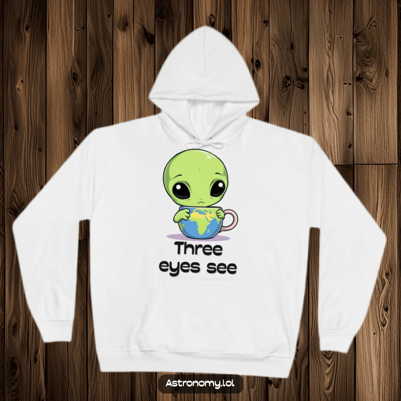 Funny alien hoodie with a confused three-eyed character fixated on an Earth mug, blending comfort with cosmic humor.